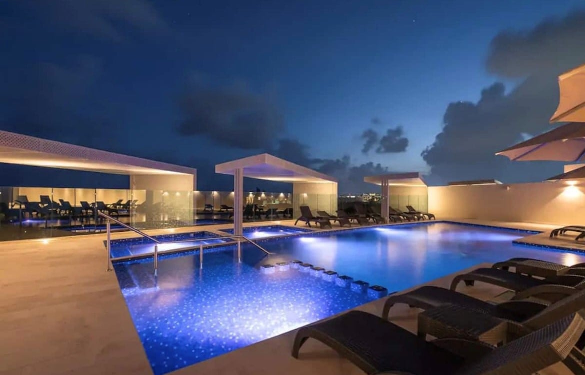 The rooftop terrace features a large pool illuminated by soft lights, surrounded by lounge chairs and umbrellas. Panoramic views of the horizon offer a captivating backdrop against the night sky, enhancing the tranquil atmosphere of the space.