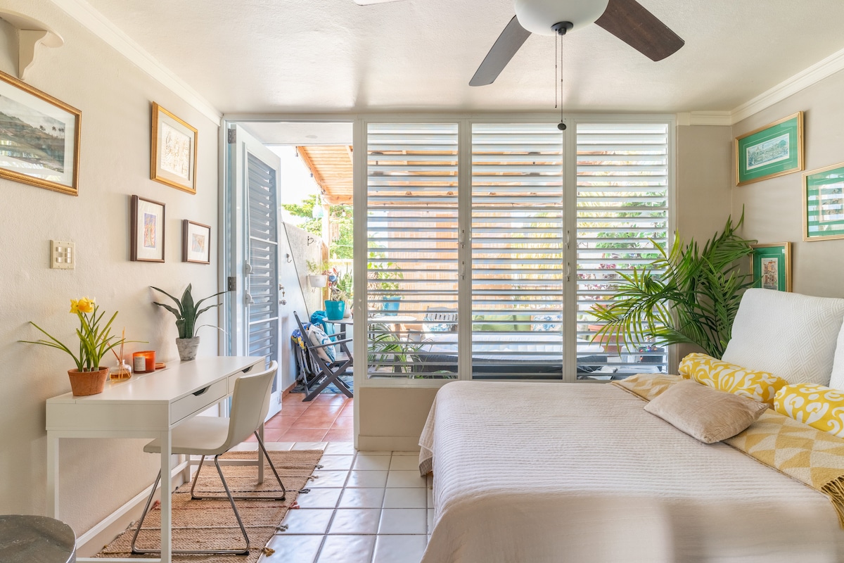 An open concept studio features a spacious bed adorned with patterned pillows. A work desk and potted plant add refreshment. Large glass windows with horizontal blinds bring natural light, while a private outdoor area is visible beyond. Earthy tones create a calming environment.