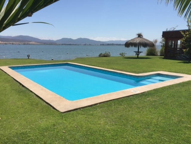 A swimming pool with clear blue water is set against a lush green lawn. A thatched-roof gazebo can be seen in the background, framed by views of the lake and distant mountains. The area is well-maintained, offering a tranquil space for relaxation.