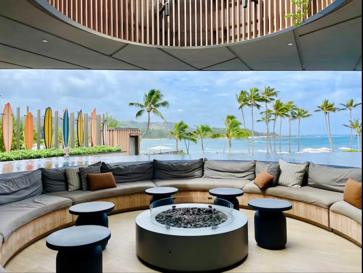 An outdoor lounging area is showcased, featuring a circular seating arrangement with soft cushions surrounding a central fire pit. Colorful surfboards line the background, and palm trees sway gently nearby, with an expansive view of the ocean and coastline visible in the distance.