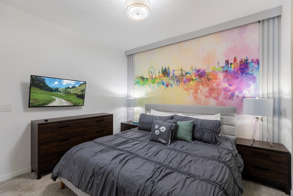 Rêvez en couleur avec cette chambre de maître moderne ! Un lit king size moelleux habillé de draps gris ardoise se trouve entre des tables de nuit élégantes surmontées de lampes à tripodes en verre. Une fresque aquarelle vibrante ancre l'espace, tandis qu'une télévision murale et une commode en grains de bois. 