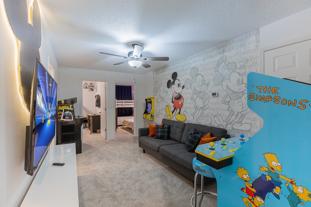 Amusez-vous en famille dans ce salon de jeux lumineux : un canapé gris confortable fait face à une télévision murale et à une arcade Simpsons classique, tandis qu'une mini armoire intégrée et une fresque Mickey rétro ajoutent une touche ludique. Tapis moelleux et ventilateur de plafond avec une ambiance chaleureuse !      