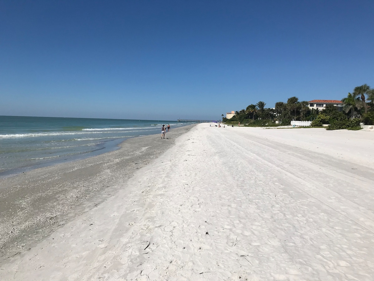 A wide expanse of soft white sand stretches along the shoreline, meeting calm turquoise waters. A few visitors are seen walking leisurely on the beach, while a pier is visible in the distance under a clear blue sky.