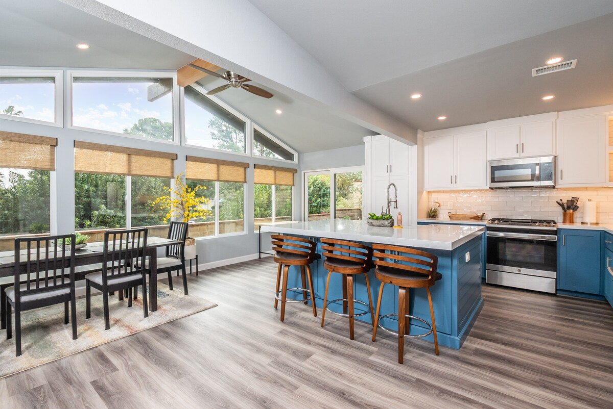 The modern kitchen features sleek white cabinetry and a spacious island with bar seating. Natural light floods the area through large windows, showcasing views of the outdoors. A dining set for four sits near the window, enhancing the inviting atmosphere.