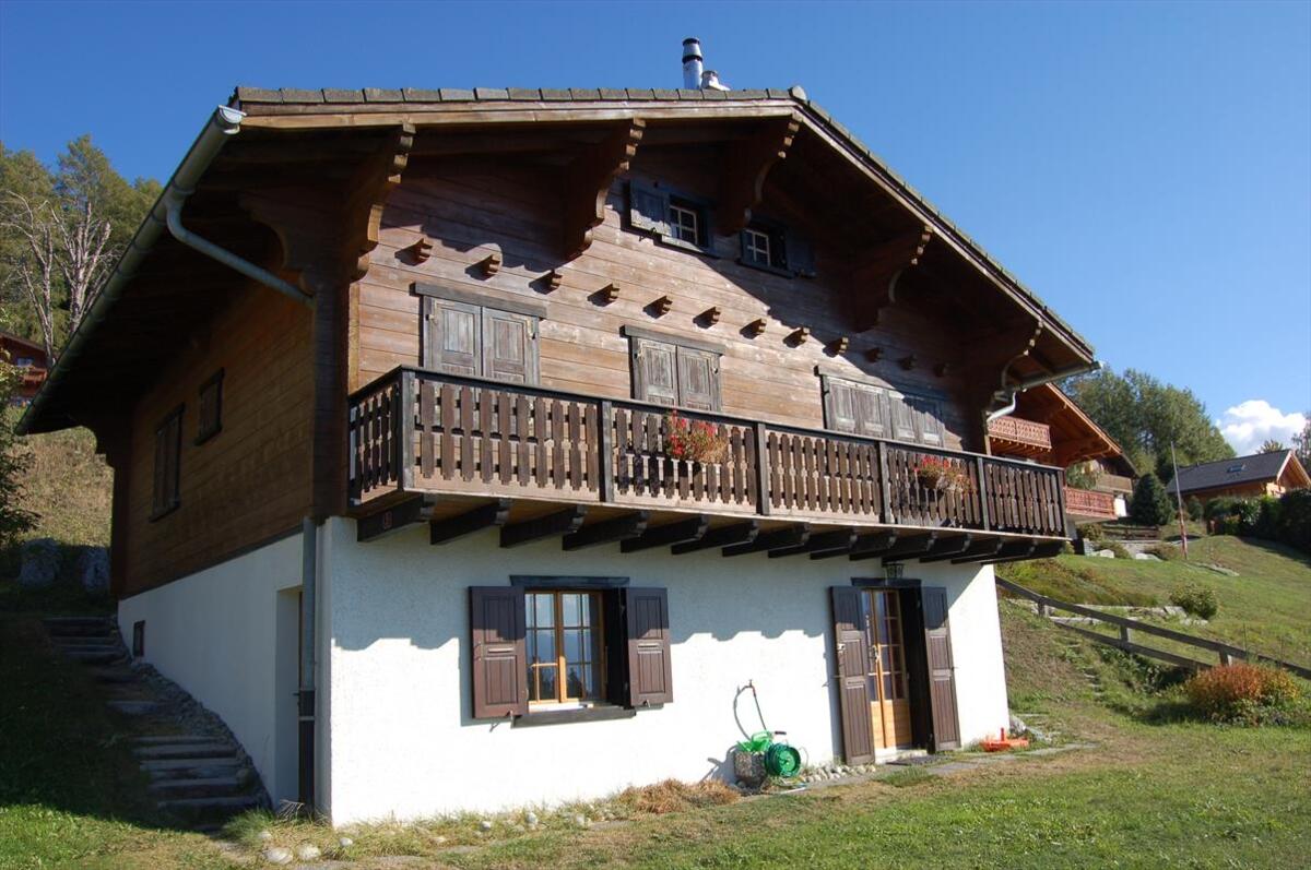 A traditional chalet with a wooden exterior features a spacious balcony adorned with flower boxes. Large windows frame the entrance, while a well-maintained lawn and gentle hillside can be seen in the background. The structure is surrounded by nature, providing a serene setting.