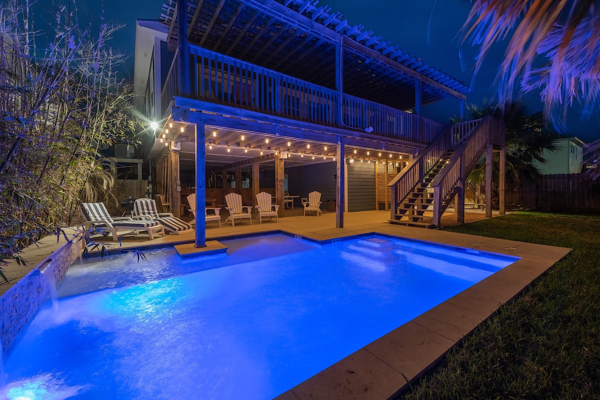A well-lit private pool area showcases soft blue waters under evening skies. Loungers line the poolside, and a covered deck features string lights, creating a relaxed outdoor space. Lush greenery surrounds the area, enhancing the overall tranquility of the setting.