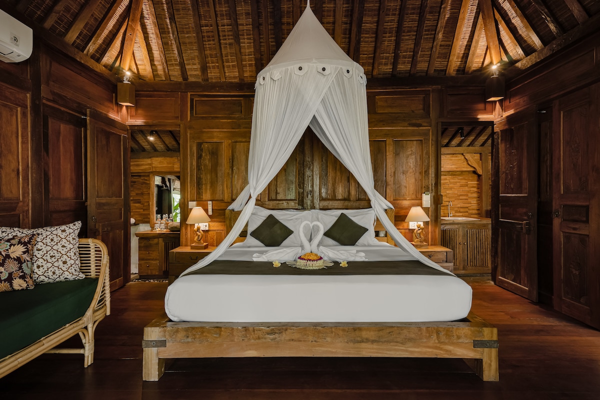 A spacious bedroom features a king-size bed draped in a white canopy, surrounded by wooden walls. Soft lighting illuminates the room, highlighting decorative elements. Two elegant lamps are positioned on nightstands, and a cozy seating area is visible to one side.
