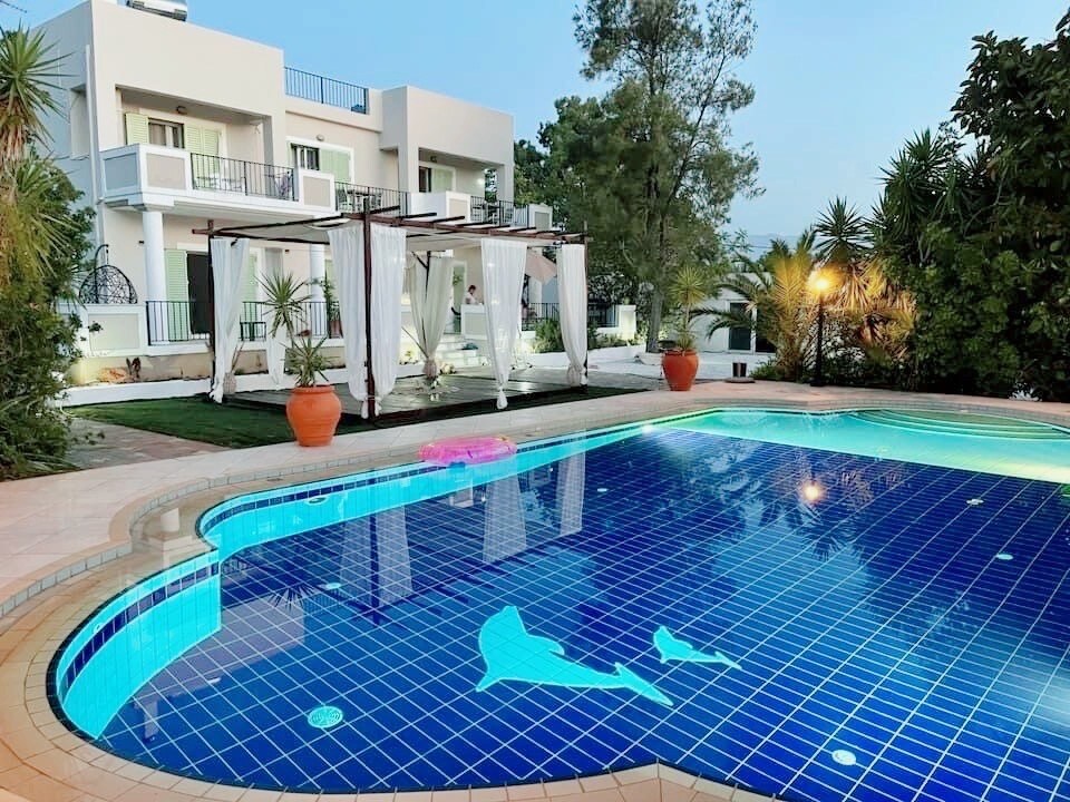 A large outdoor pool is surrounded by lush greenery and palm trees, featuring unique tile designs. A shaded seating area with draped fabric is visible, creating a tranquil space. The villa's modern architecture stands in the background, contributing to the serene ambiance.