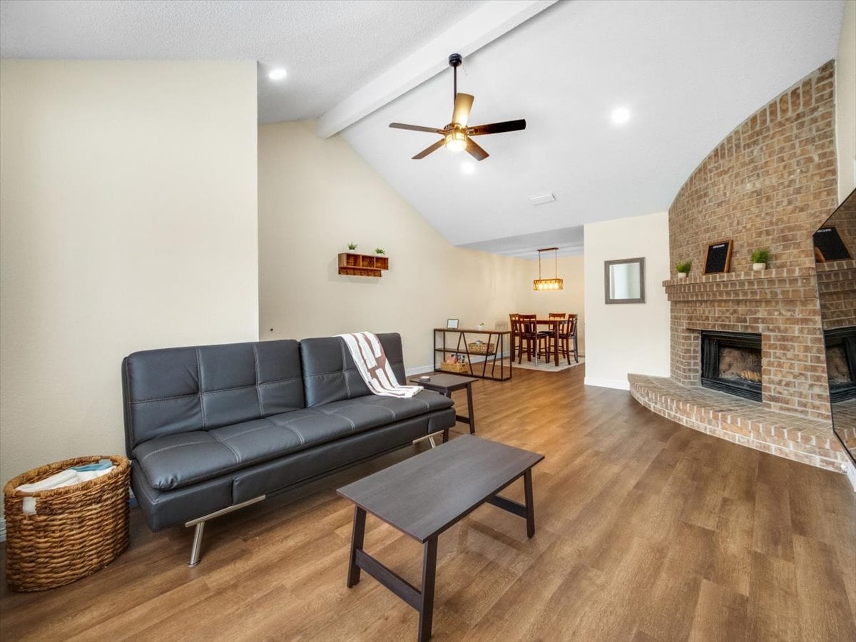 A spacious living area features a dark leather couch complemented by a wooden coffee table. A brick fireplace provides warmth, and a ceiling fan circulates air. In the background, a dining table set for six is visible, adding to the inviting ambiance.