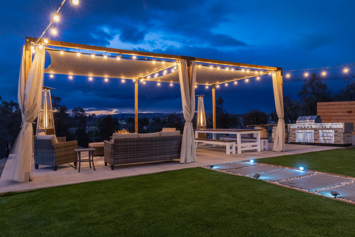An outdoor patio is illuminated by string lights, creating a warm atmosphere. Comfortable seating is arranged underneath a large pergola, complemented by a dining table. A professional grill is visible to one side, with a well-maintained lawn extending around the area.