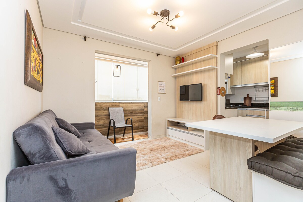 A well-lit living area features a cozy gray sofa positioned near a wooden shelving unit and a small television. A dining table is situated adjacent to the kitchen. Large windows provide natural light, and a light-colored rug adds warmth to the space.
