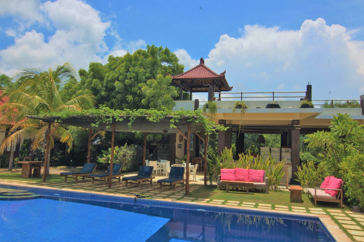 The outdoor area features a large swimming pool surrounded by lounge chairs, with bright pink and deep blue seating arranged under shaded pergolas. A traditional gazebo is visible above, and lush greenery enhances the tranquil atmosphere of the garden space.