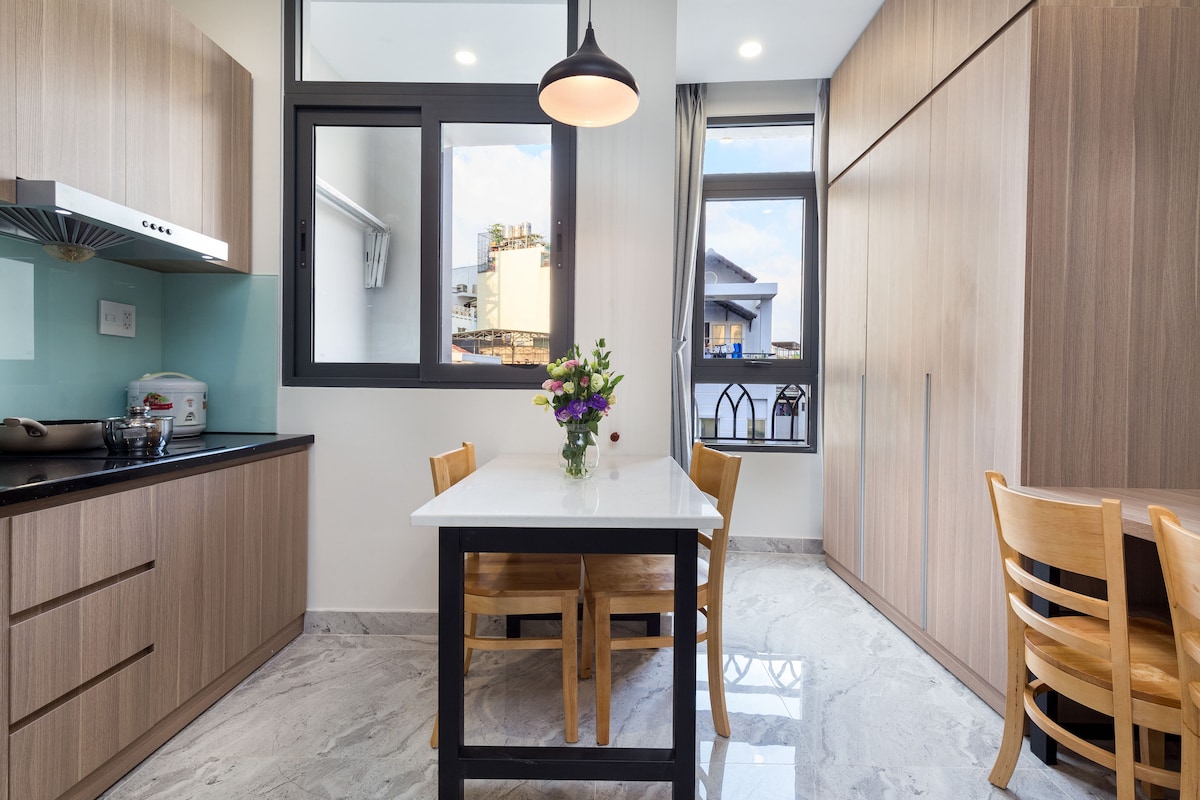A light-filled kitchen area is observed, featuring a modern table with four wooden chairs. Sleek cabinetry lines the walls, and a small bouquet of flowers serves as a centerpiece. Two large windows provide an airy view, enhancing the spaciousness of the room.