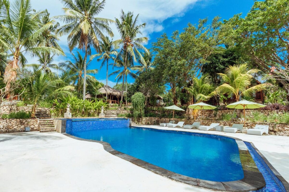 A vast swimming pool is surrounded by lush greenery and palm trees. Comfortable lounge chairs are positioned under umbrellas, offering shaded seating. The pool's clear blue water reflects the sunny sky, creating a serene outdoor retreat.