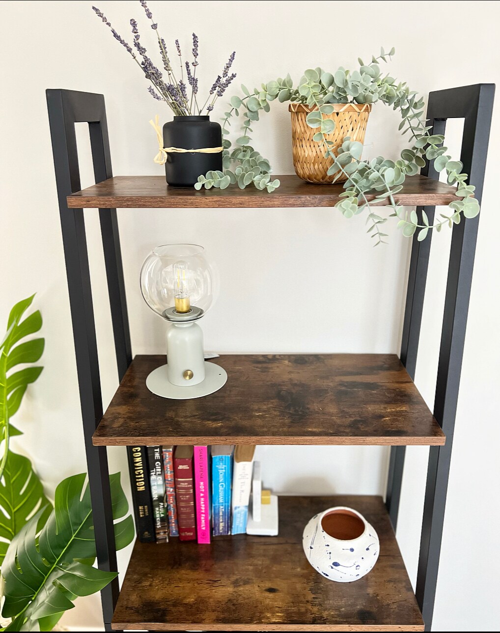 A wooden bookshelf with a black metal frame displays multiple decorative items. A potted plant sits on the top shelf next to a small candle, while lavender and eucalyptus accents provide natural elements. Books are organized on the lower shelves, with a decorative vase nearby.