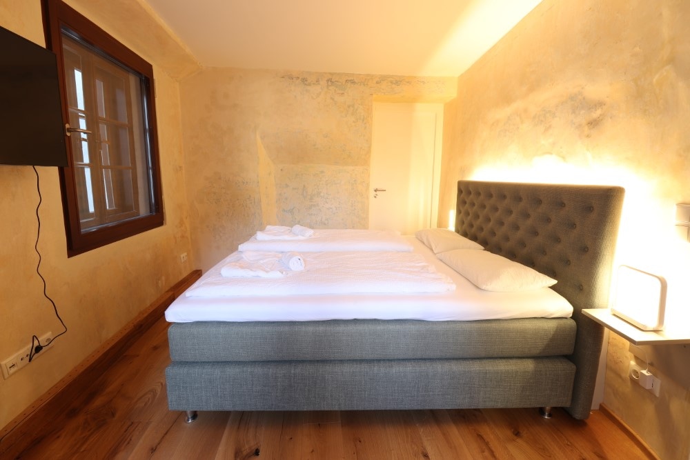 A well-furnished sleeping area features a spacious bed with a light-coloured duvet and pillows. The walls have a warm texture, and a flat-screen TV is mounted on one side. Natural light enters through a small window, with light wood flooring enhancing the ambiance.