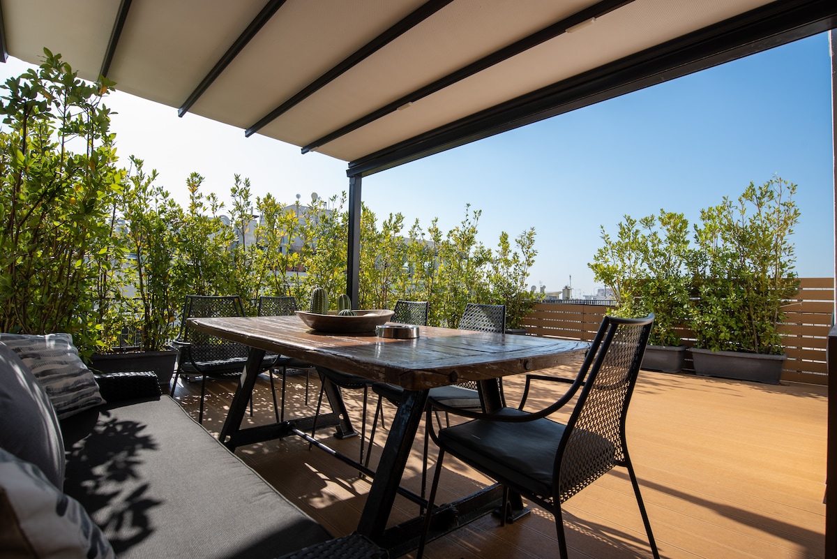 A private balcony is featured with a large dining table surrounded by black metal chairs. Comfortable seating is provided on a dark sofa, while greenery enhances the space, creating a serene outdoor environment. The sun protection awning adds convenience for enjoyment throughout the day.