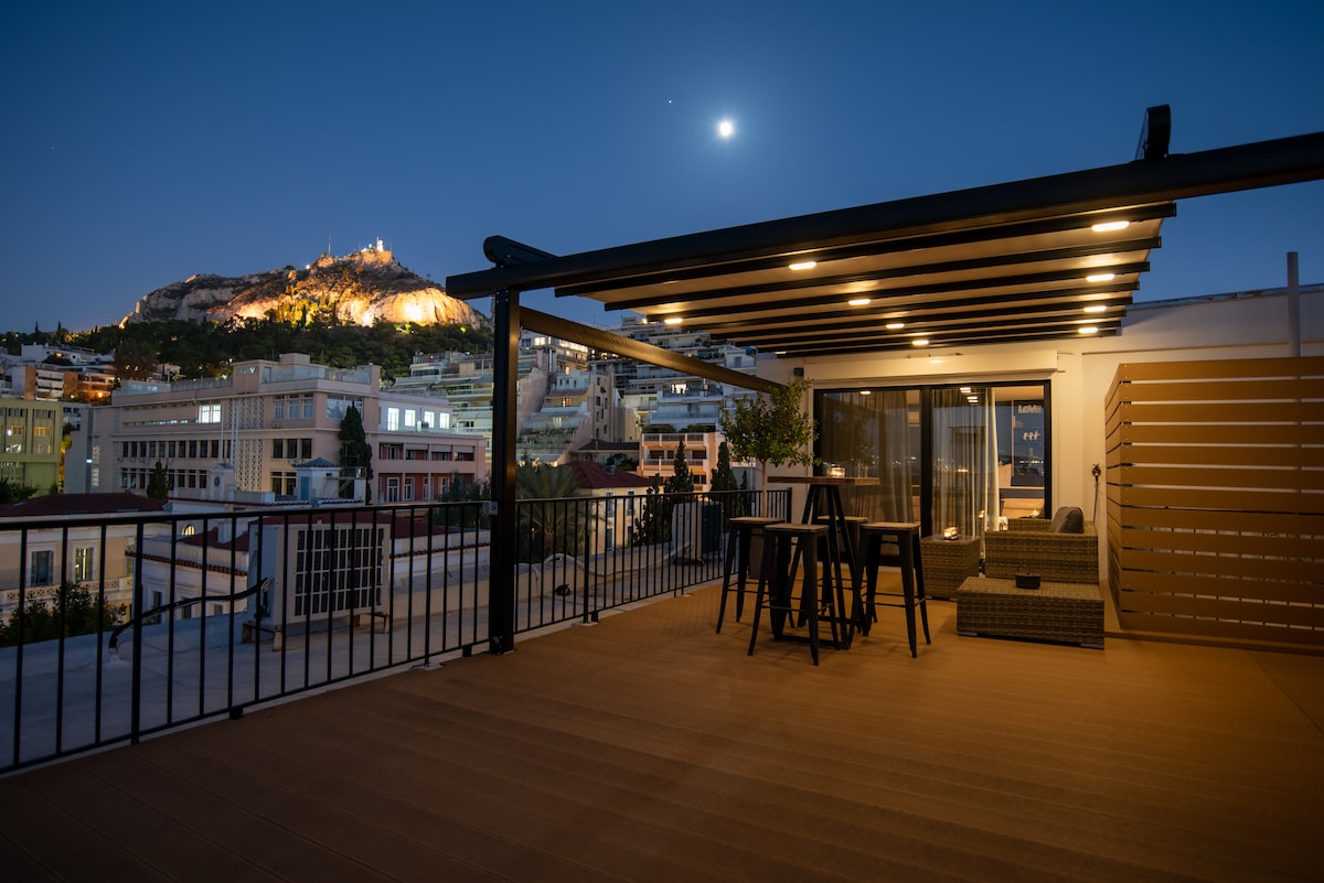 An evening view of a private balcony featuring a dining table and chairs, accompanied by a cozy seating area. The Acropolis is illuminated in the background, and a full moon casts a gentle glow over the scene, highlighting the balcony's inviting atmosphere.