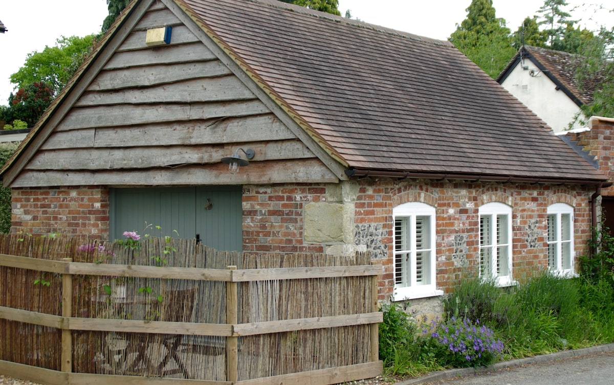 The exterior of a quaint studio cottage is displayed, featuring a mix of brick and wooden paneling. A wooden fence surrounds the front yard, which is adorned with various flowers and plants. White window shutters enhance the cottage's charm, with an inviting entryway visible.