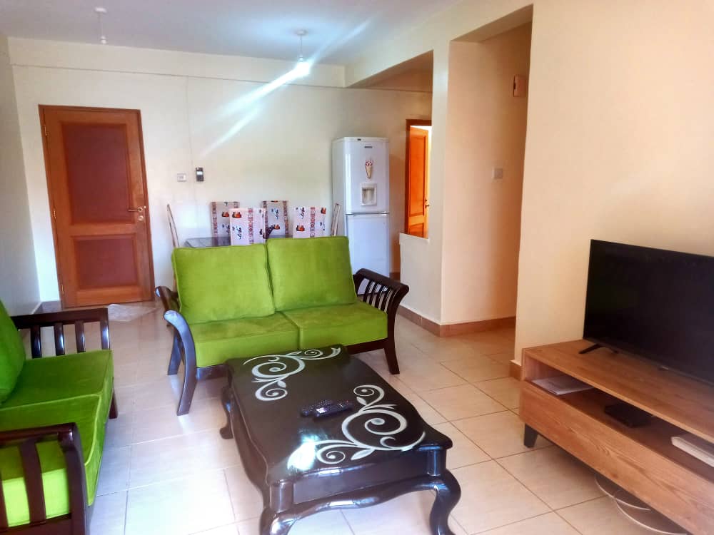 A living space is depicted with two green sofas arranged around a wooden coffee table featuring intricate detailing. A television sits on a wooden stand, while a refrigerator is visible in the background, showing a functional and comfortable environment for relaxation.