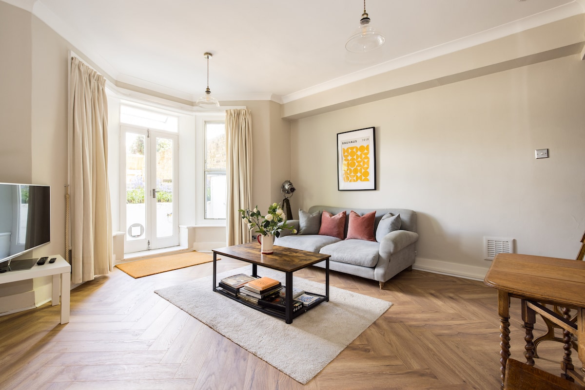 A light-filled living room features large windows dressed with flowing curtains. A comfortable grey sofa is positioned alongside a wooden coffee table, adorned with a decorative plant. A warm area rug complements the wooden floor, while a stylish dining table adds to the inviting atmosphere.