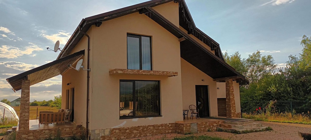 The exterior of a modern structure is shown, featuring a combination of stone and stucco. Large windows allow natural light into the building. A covered porch area is visible, accompanied by a single chair. Lush greenery surrounds the house, enhancing the serene setting.