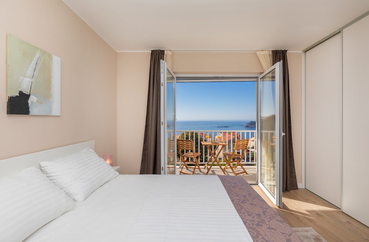 A bright bedroom is furnished with a spacious white bed, featuring crisp linens. Large glass doors open to a balcony showcasing panoramic views of the Adriatic Sea. A small outdoor dining set is visible, enhancing the serene coastal ambiance.