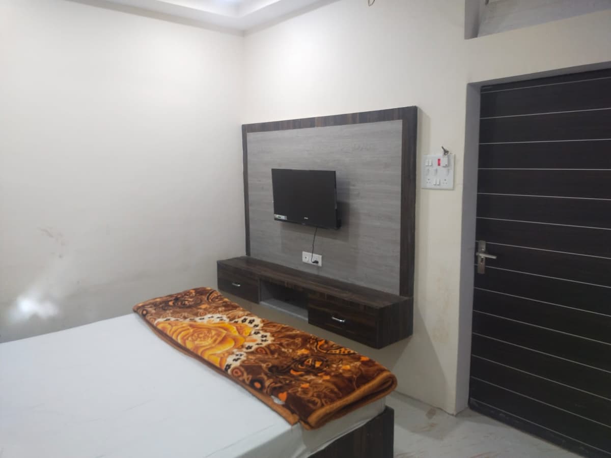A simple sleeping area features a spacious bed with a patterned blanket draped over it. A wall-mounted TV is positioned above a dark wooden console, while a door with a modern design is visible to the right.