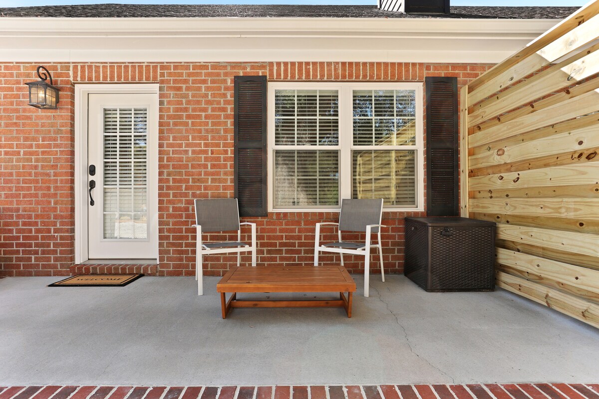 A private outdoor patio features a small wooden coffee table accompanied by two white chairs. The brick wall and wooden fence create a cozy atmosphere, while a door with a glass panel provides access to the interior space.
