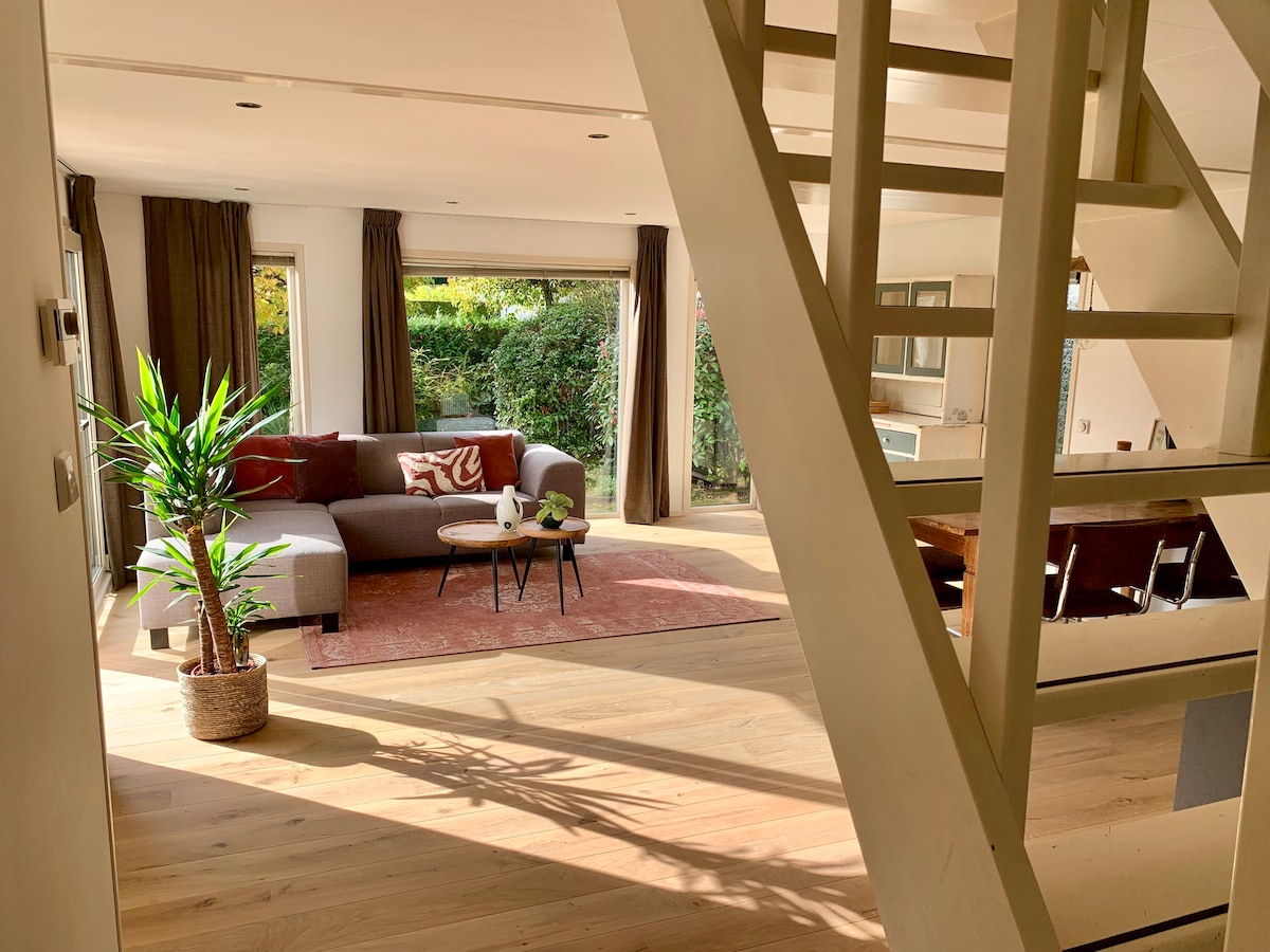 A spacious living area is filled with natural light, showcasing a comfortable gray sofa accented with cushions. A potted plant adds a touch of greenery, while a soft area rug complements the wooden flooring. Large windows provide views of the lush surroundings.