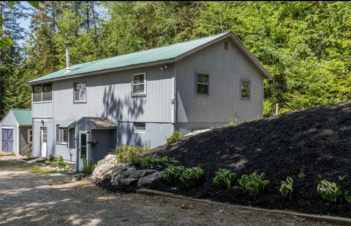 The exterior of a two-story house is depicted, featuring a grey siding and a green metal roof. Surrounded by lush greenery, a gravel driveway leads to the entrance, while a small garden with hostas is visible in the foreground.