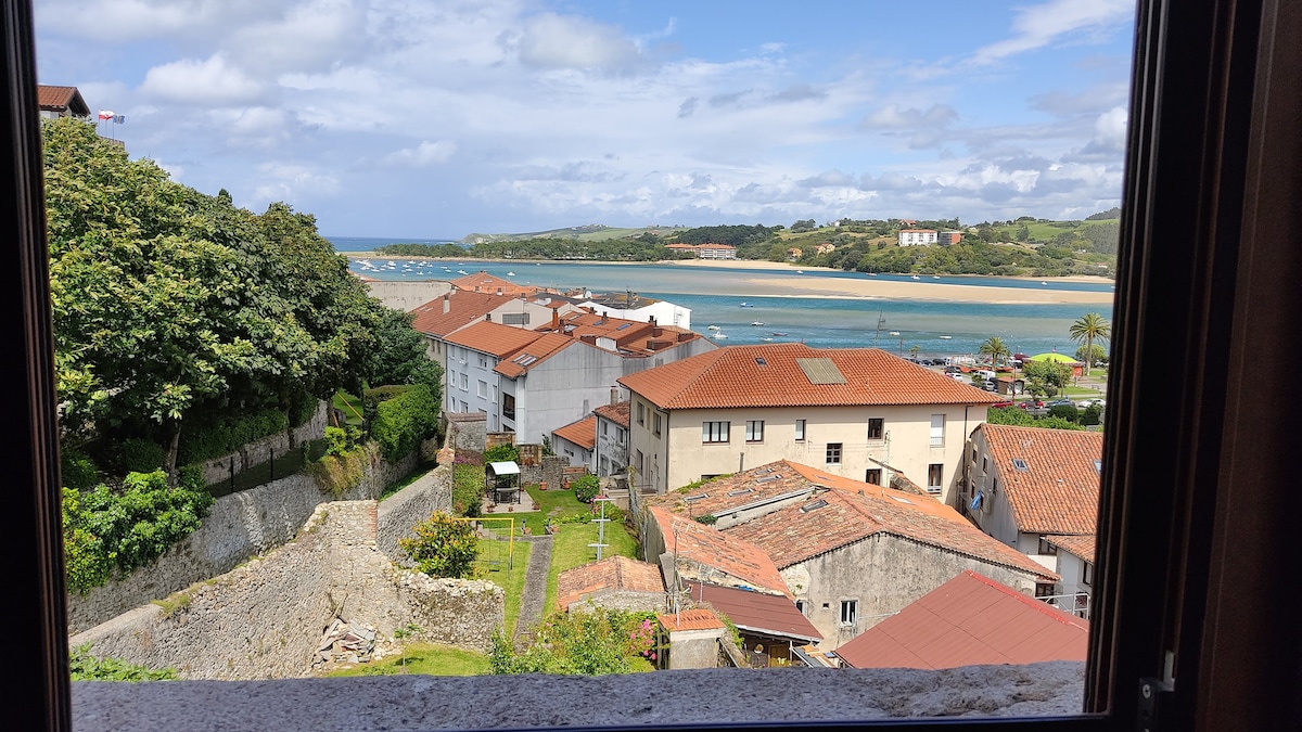 A view from a window showcases a serene landscape, including rooftops of nearby buildings with red-tiled roofs and lush greenery. The distant coastline features sandy shores and gently rolling hills under a partly cloudy sky.