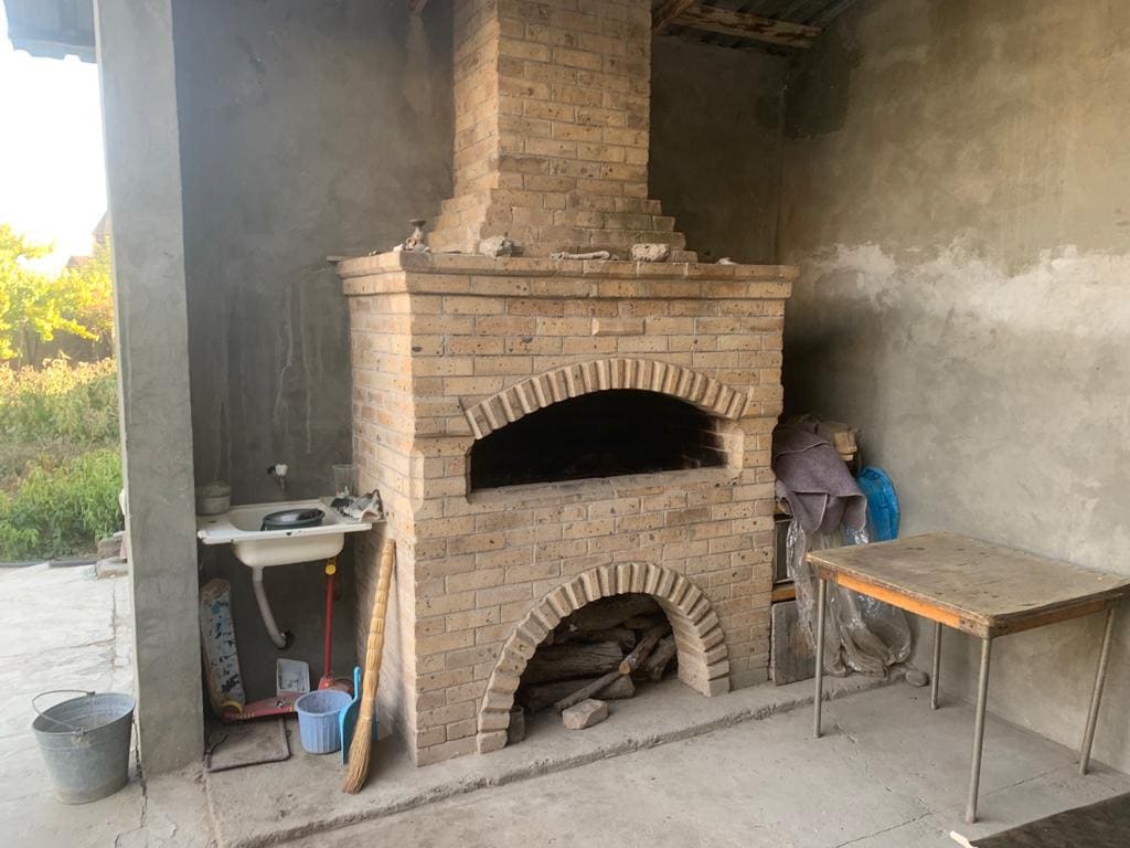 A brick oven is positioned against an unfinished concrete wall, featuring two distinct arches for cooking. Nearby, a small sink and a table are visible, along with basic cleaning supplies. Natural light filters in from an unseen window, illuminating the rustic space.
