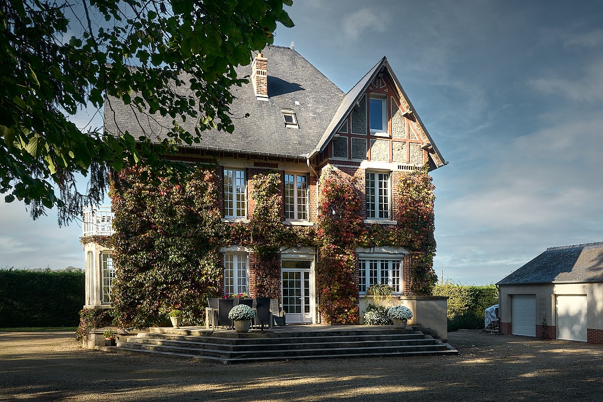 A charming three-story house features a combination of brick and timber elements, enveloped by climbing greenery. A set of stairs leads to a welcoming terrace with outdoor seating, offering a view of the landscaped surroundings under a cloudy sky.