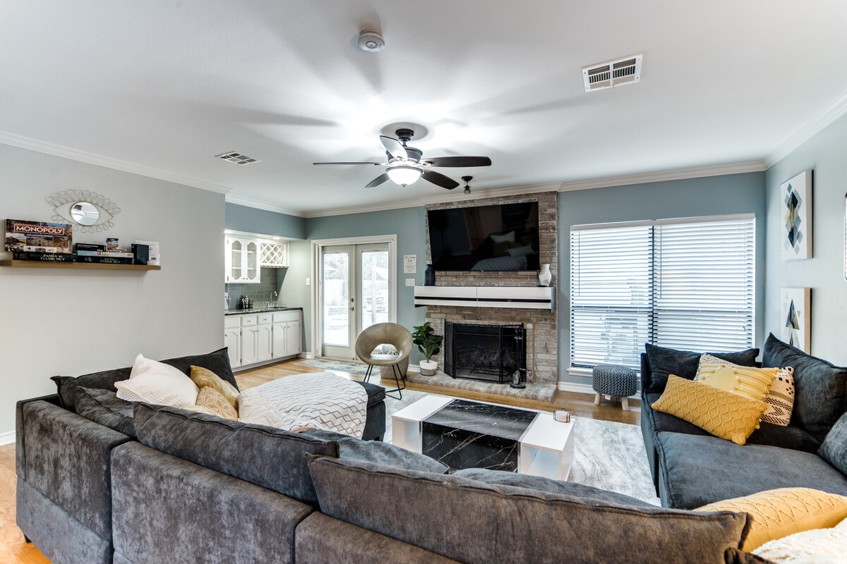 A spacious living area is decorated with a large sectional sofa featuring multiple throw pillows. A coffee table sits in the center, and a fireplace is positioned beneath a mounted television. Natural light enters through large windows, illuminating the neutral color palette.