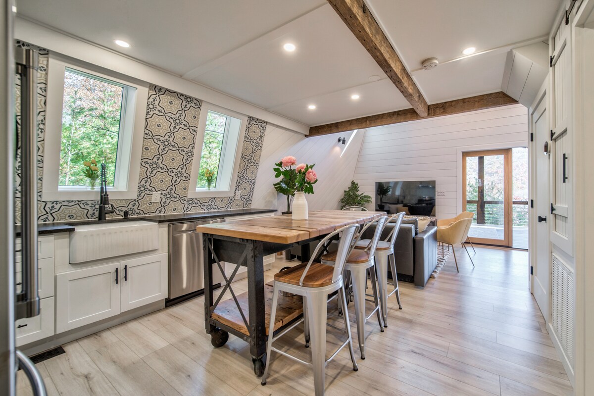 The kitchen area features a large wooden dining table surrounded by metal and wooden chairs. Tile accents on the walls provide a stylish backdrop to the space, while the large windows allow natural light to brighten the room. A cozy seating area with a television is visible near glass doors that lead outdoors.