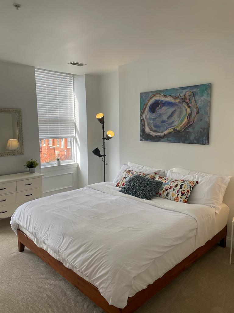 A spacious bedroom is illuminated by natural light streaming through a large window. The bed features a soft white comforter with decorative pillows that add a touch of color. A tall lamp stands beside the bed, and a dresser with a mirror is positioned against the wall.