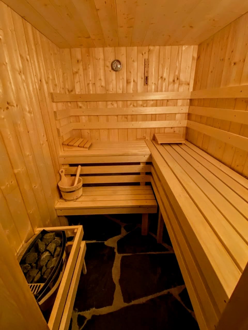 A wooden sauna features simple benches along the walls and a stone floor, enhancing a sense of warmth. A sauna stove with rocks is visible, along with a traditional wooden bucket and ladle. Natural wood paneling creates a calming environment.