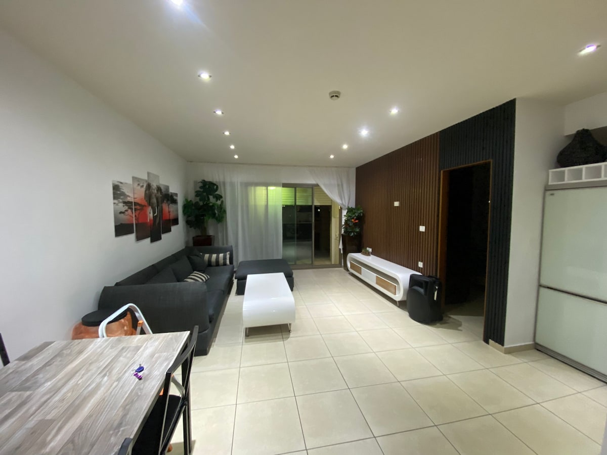 A spacious living area features a comfortable gray sofa and a sleek white coffee table. A dining table with wooden elements is positioned nearby. Large sliding doors open to a terrace, framed by soft curtains, while a fridge and decorative plants add functional and aesthetic touches.