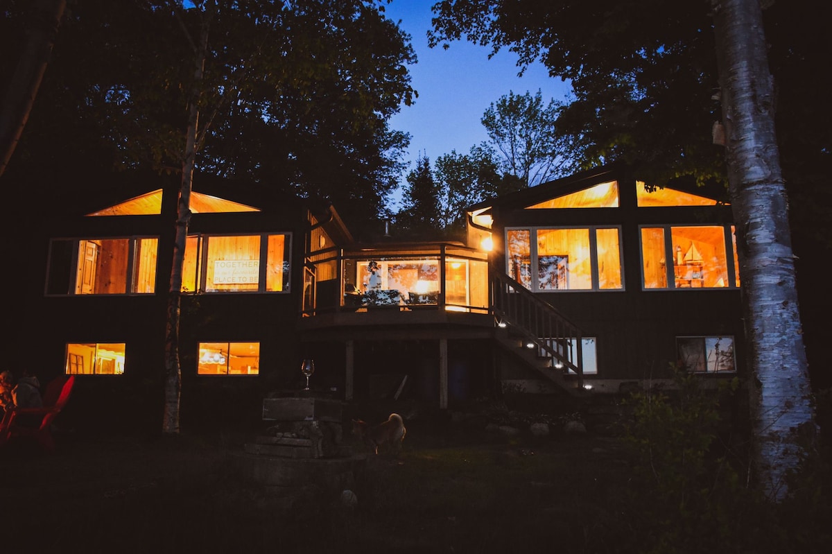 The cottage is depicted at dusk, showcasing warm light emanating from expansive windows. The structure features multiple gabled roofs and a spacious deck accessed by a staircase. Surrounded by trees, a fire pit is located in the foreground, enhancing the serene outdoor setting.