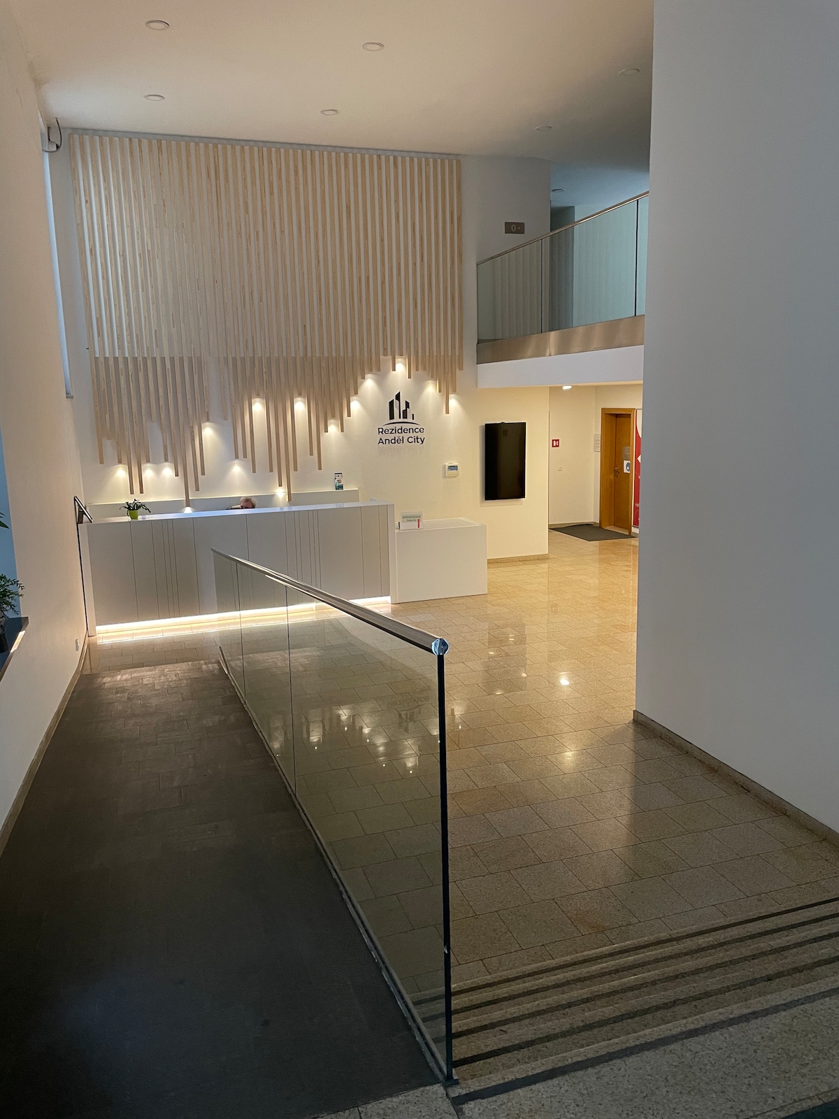 The reception area features a modern design with wood paneling and soft lighting. A sleek front desk is visible, accompanied by a minimalist decor. A glass railing provides safety near the staircase leading to the upper level, enhancing the open, airy atmosphere of the space.