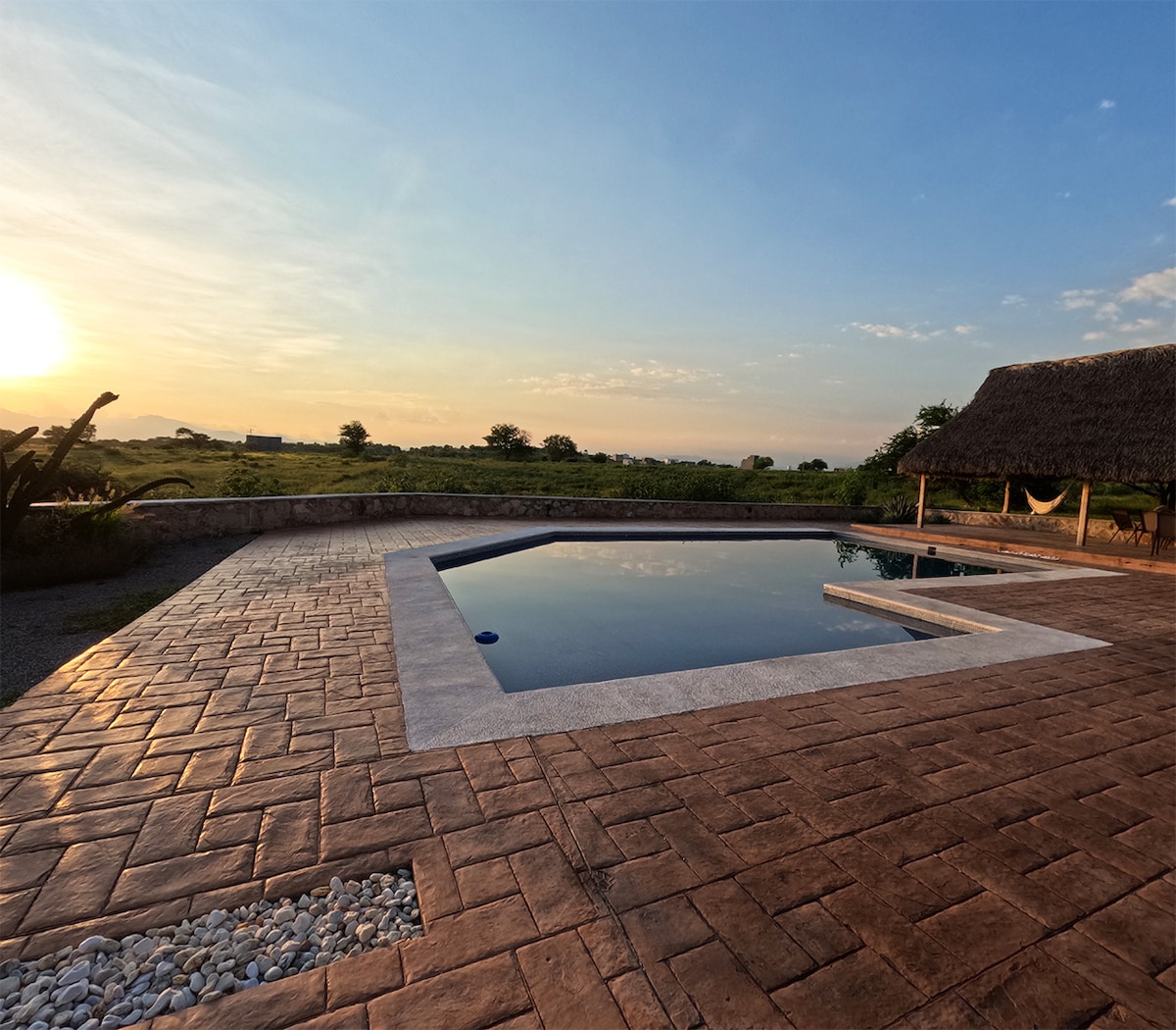 A spacious pool area is set against a scenic landscape, featuring a rectangular pool with a sun ledge and a gently sloping deck. A straw palapa provides shade nearby, while a soft glow from the sunset enhances the natural surroundings.