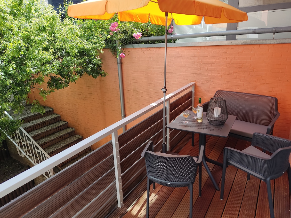 A sunlit terrace is highlighted by an orange umbrella providing shade over a small table and chairs. Lush greenery and flowering plants create a vibrant backdrop. A set of stairs leads down from the terrace to the lower level, enhancing the outdoor space.
