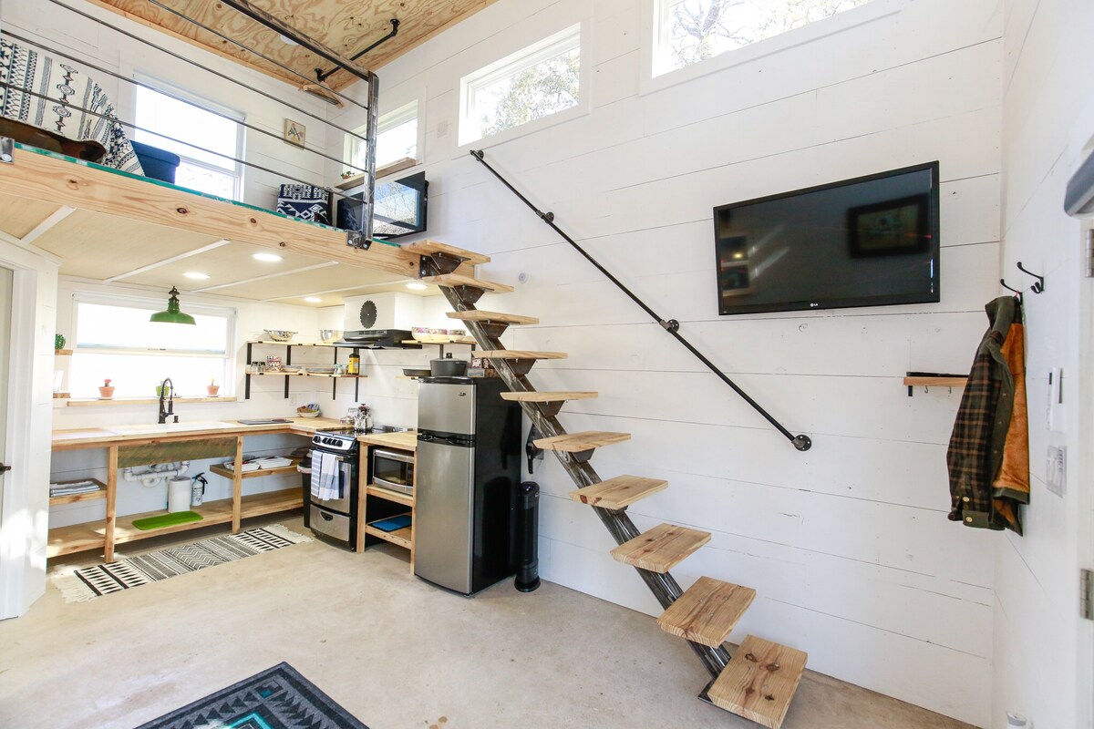 A modern interior showcases a compact kitchen with butcher block countertops and stainless steel appliances. A wooden staircase leads to a lofted area. Natural light floods the space through large windows, highlighting the minimalist design and open layout.