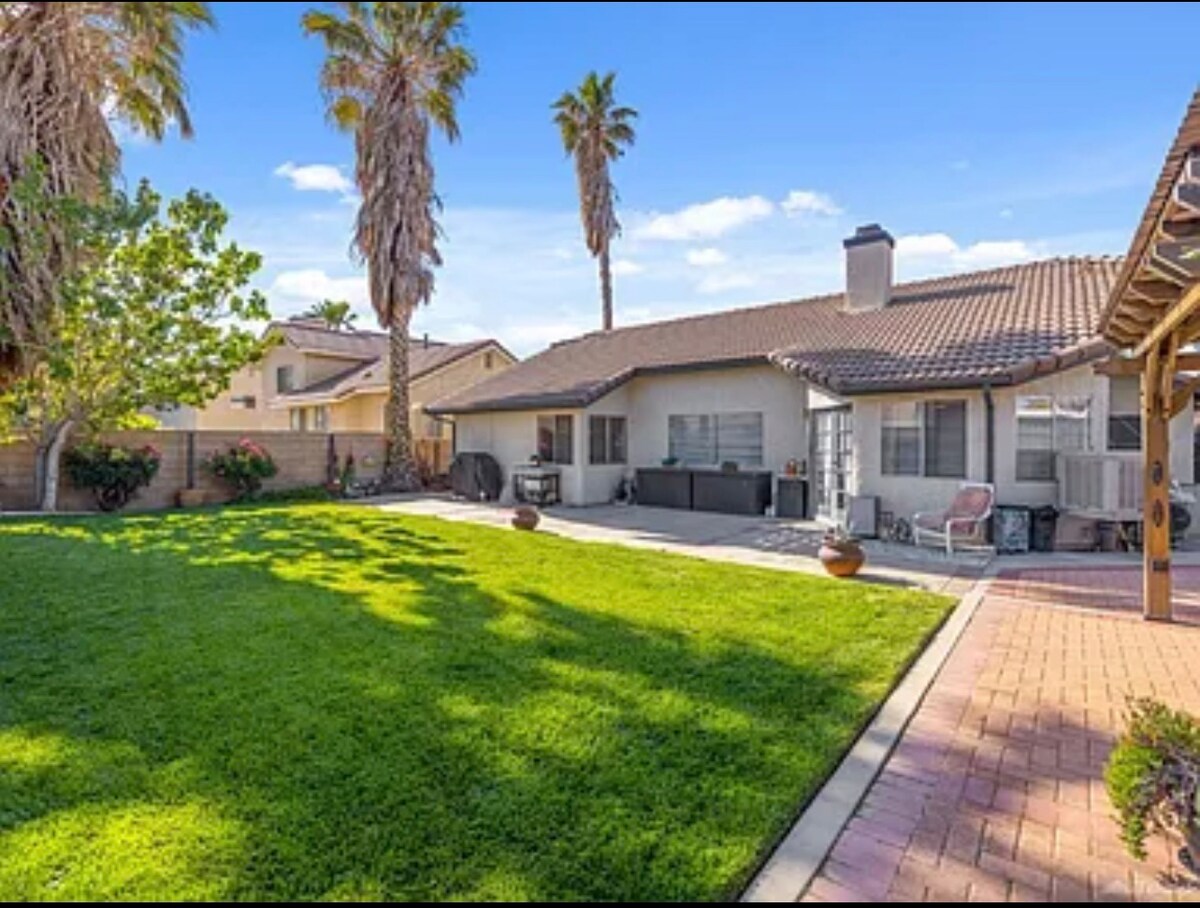 A spacious outdoor area is framed by tall palm trees and features a well-maintained lawn. The patio includes a shaded seating area with comfortable furniture, providing a relaxing space for guests. A clear blue sky enhances the inviting atmosphere.