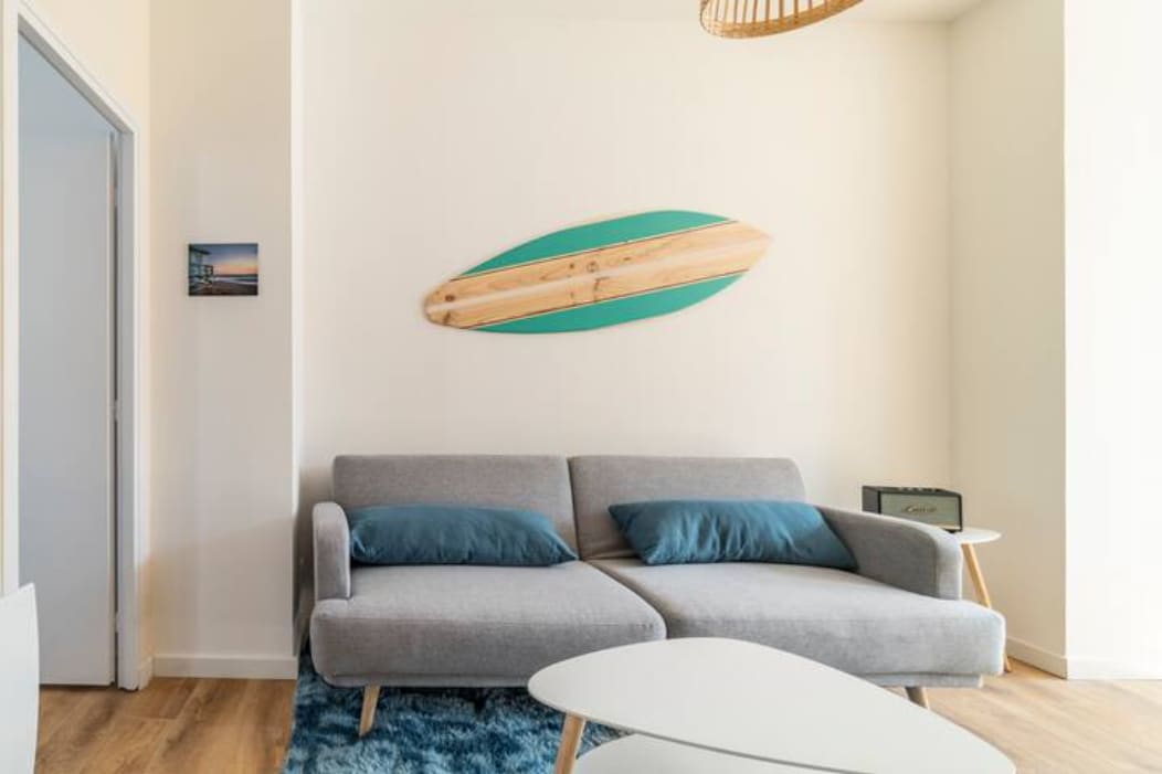 A modern living area features a light gray sofa with teal cushions, complemented by a wooden surfboard mounted on the wall. A soft rug enhances the space, while minimalist tables provide clean lines and functionality, contributing to a serene atmosphere.