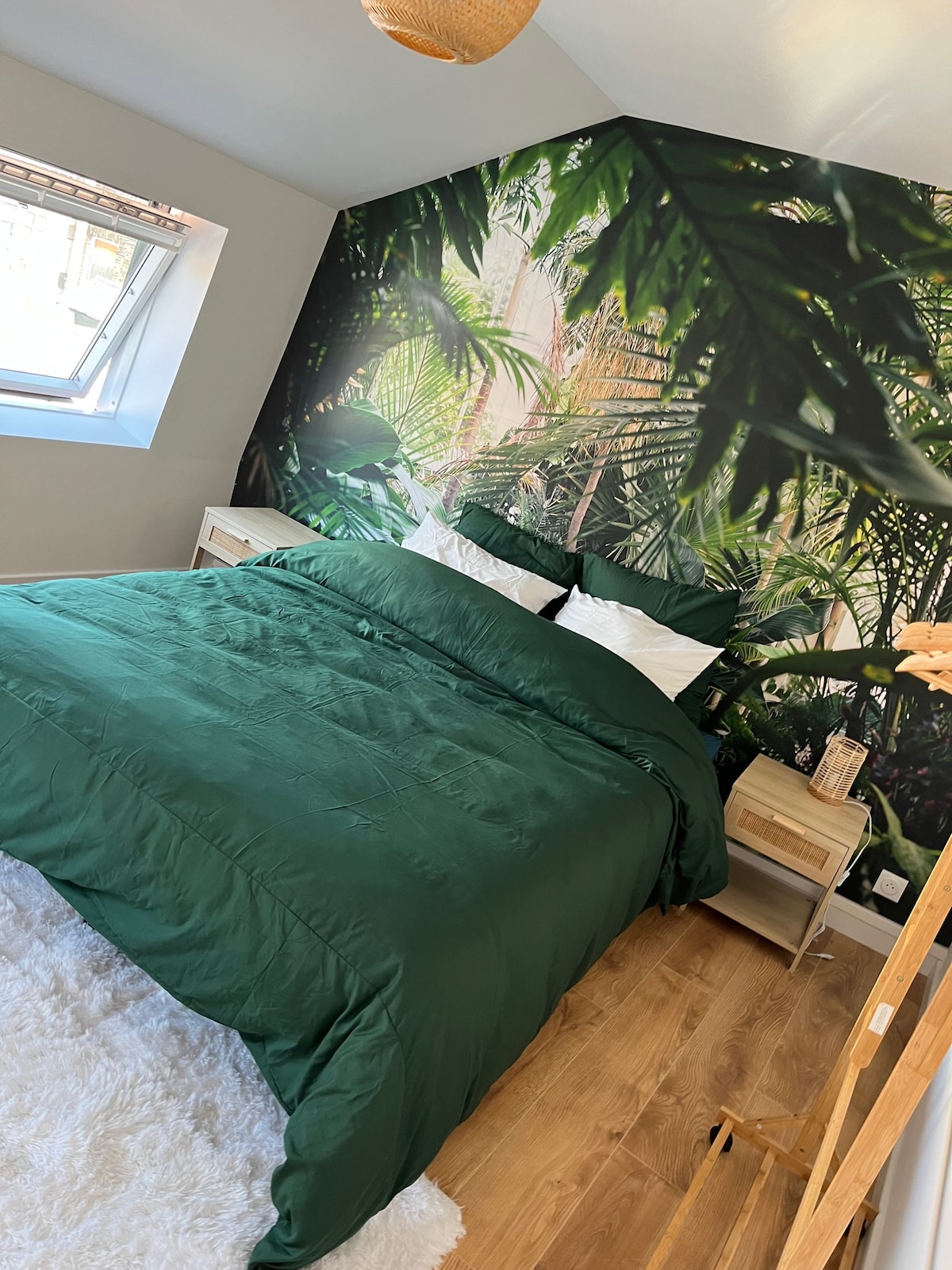 A tropical-themed bedroom features green bedding that complements a leafy wall mural. Natural light filters in through a slanted window, creating a bright and airy ambiance. Nightstands with lamps are placed on either side of the bed, enhancing the room's serene vibe.
