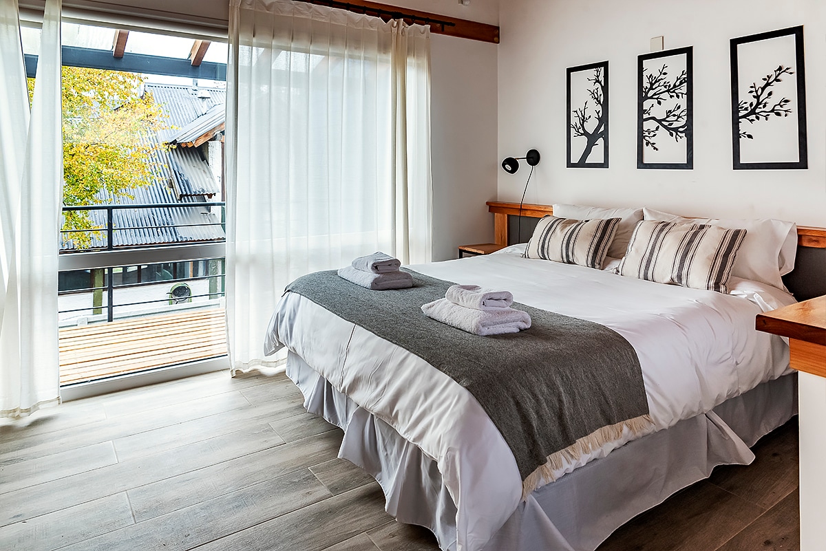 A bright bedroom features a spacious bed with grey and white linens, complemented by two neatly placed towels. Natural light filters through sheer curtains, illuminating framed artwork on the walls. A sliding glass door opens to a wooden balcony, offering views of the outdoor area.