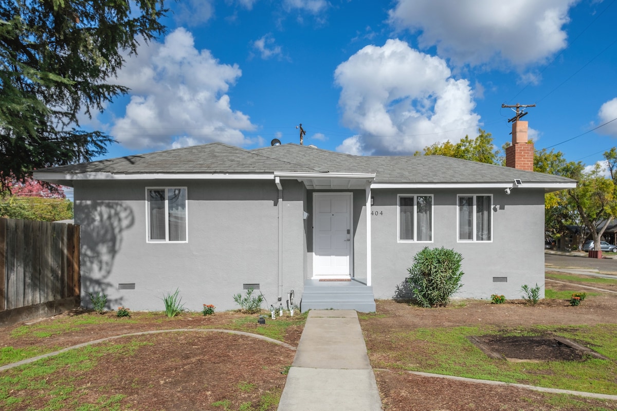 A single-story home with a light gray exterior is presented, featuring a central front door framed by two windows. A concrete pathway leads to the entrance, surrounded by neatly maintained landscaping, including patches of grass and small shrubs. Fluffy clouds drift in a bright blue sky.