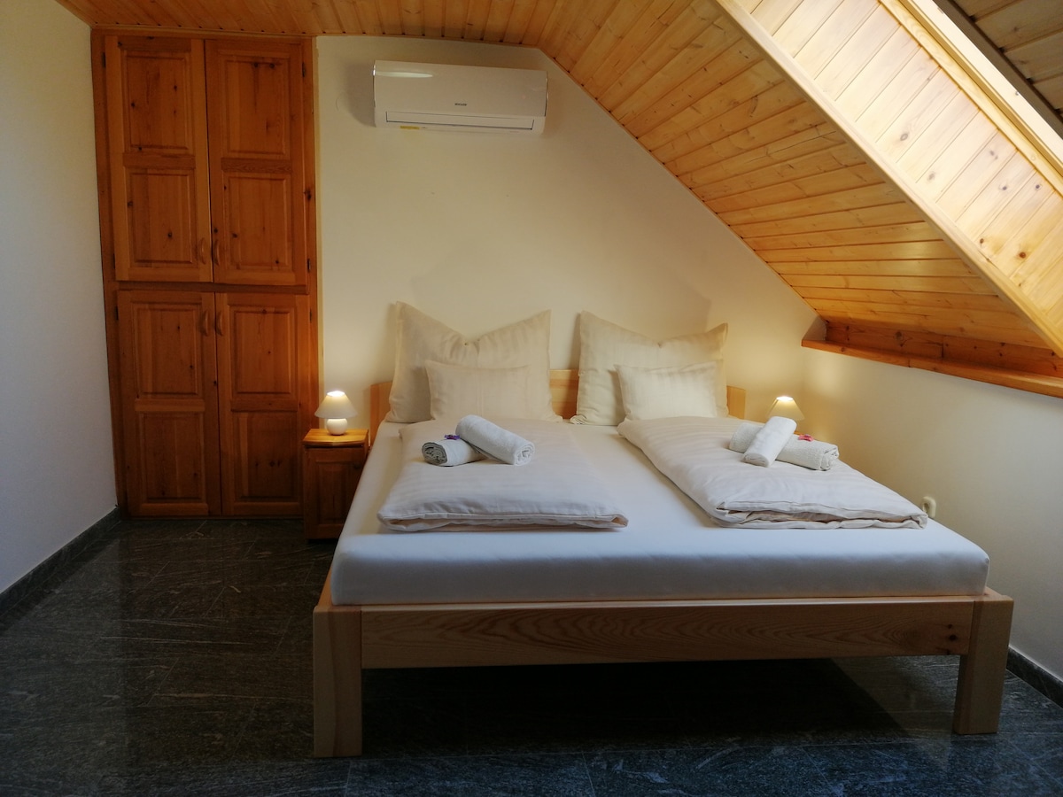 A comfortable bedroom is showcased with two spacious beds placed side by side, adorned with soft linens and rolled towels. Wooden furniture, including a wardrobe and bedside tables, complements the warm wood paneling of the slanted ceiling, while soft lighting adds a relaxing ambiance.